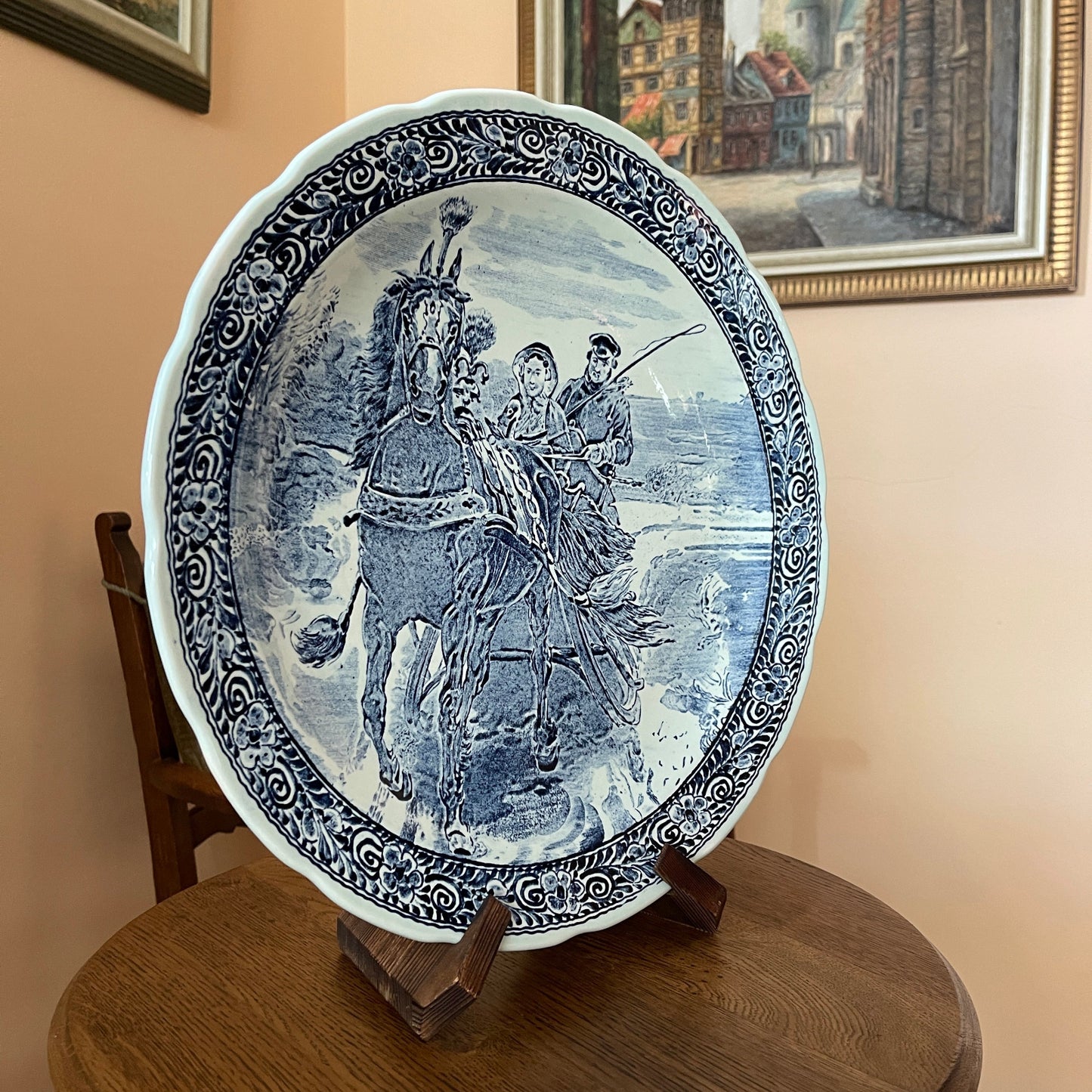 Decorative plate with blue and white design of people on horseback, placed on a wooden stand.