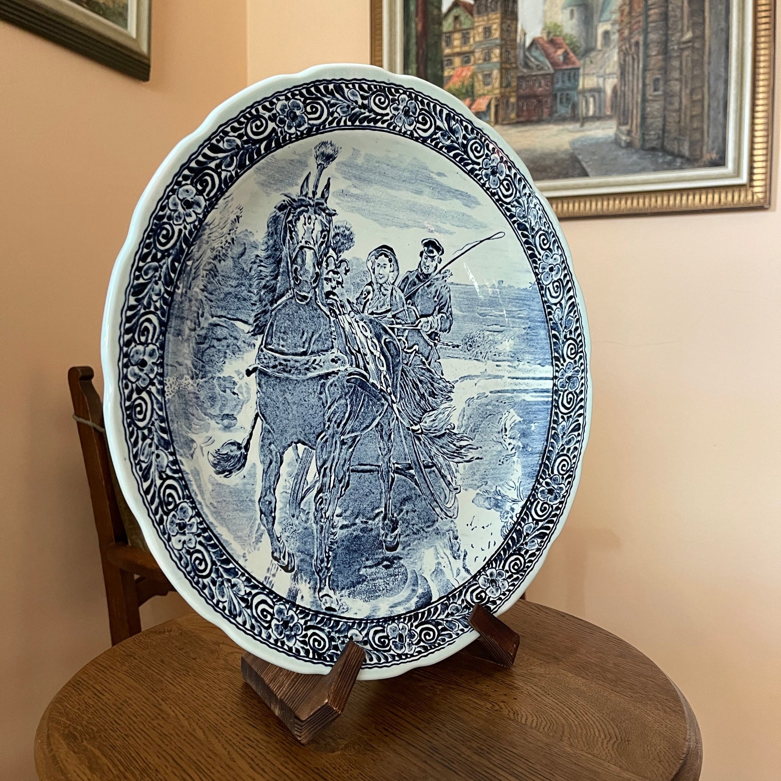Decorative plate with blue and white design of people on horseback, placed on a wooden stand.