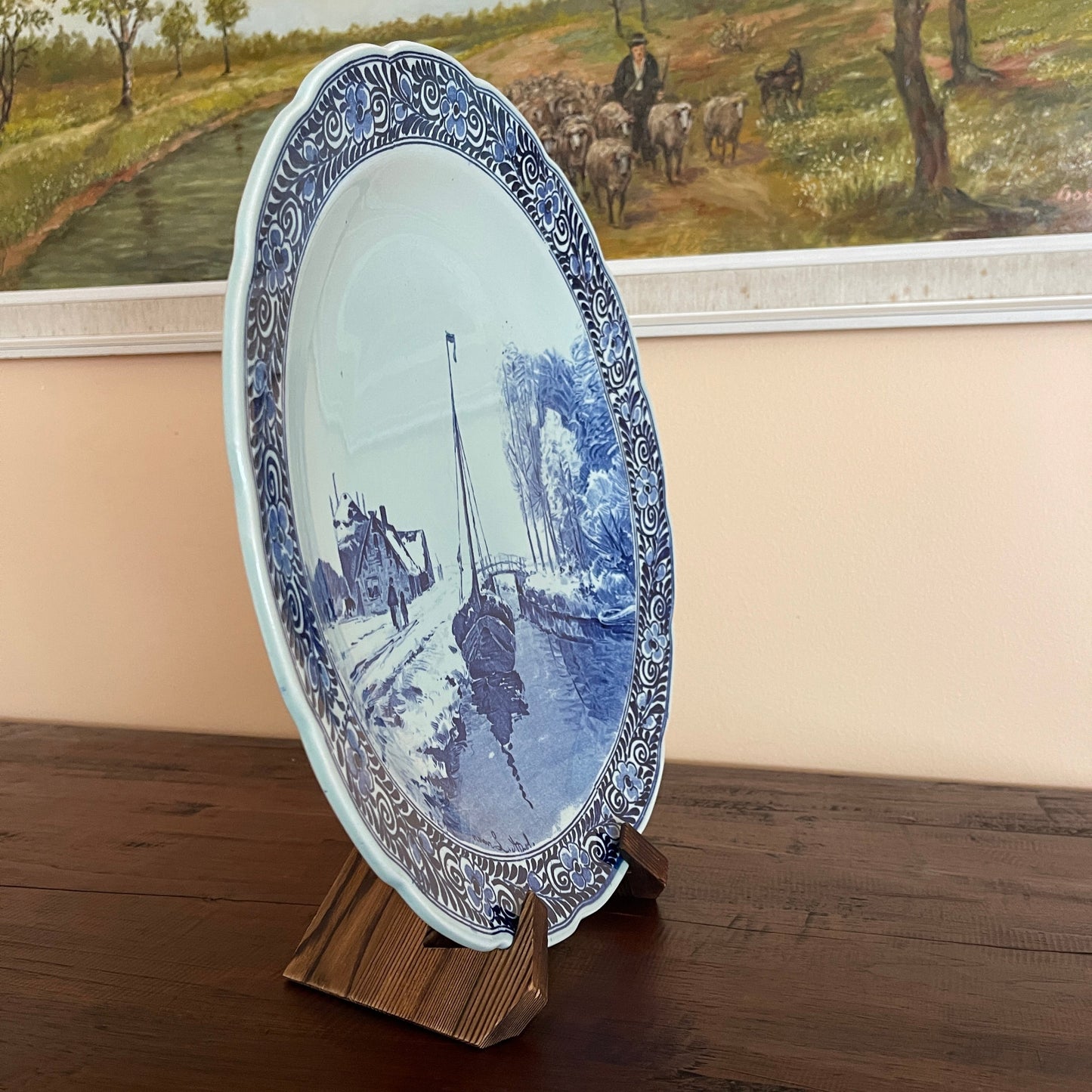 Decorative plate with blue and white design on a wooden stand, with a painting of a pastoral scene in the background.