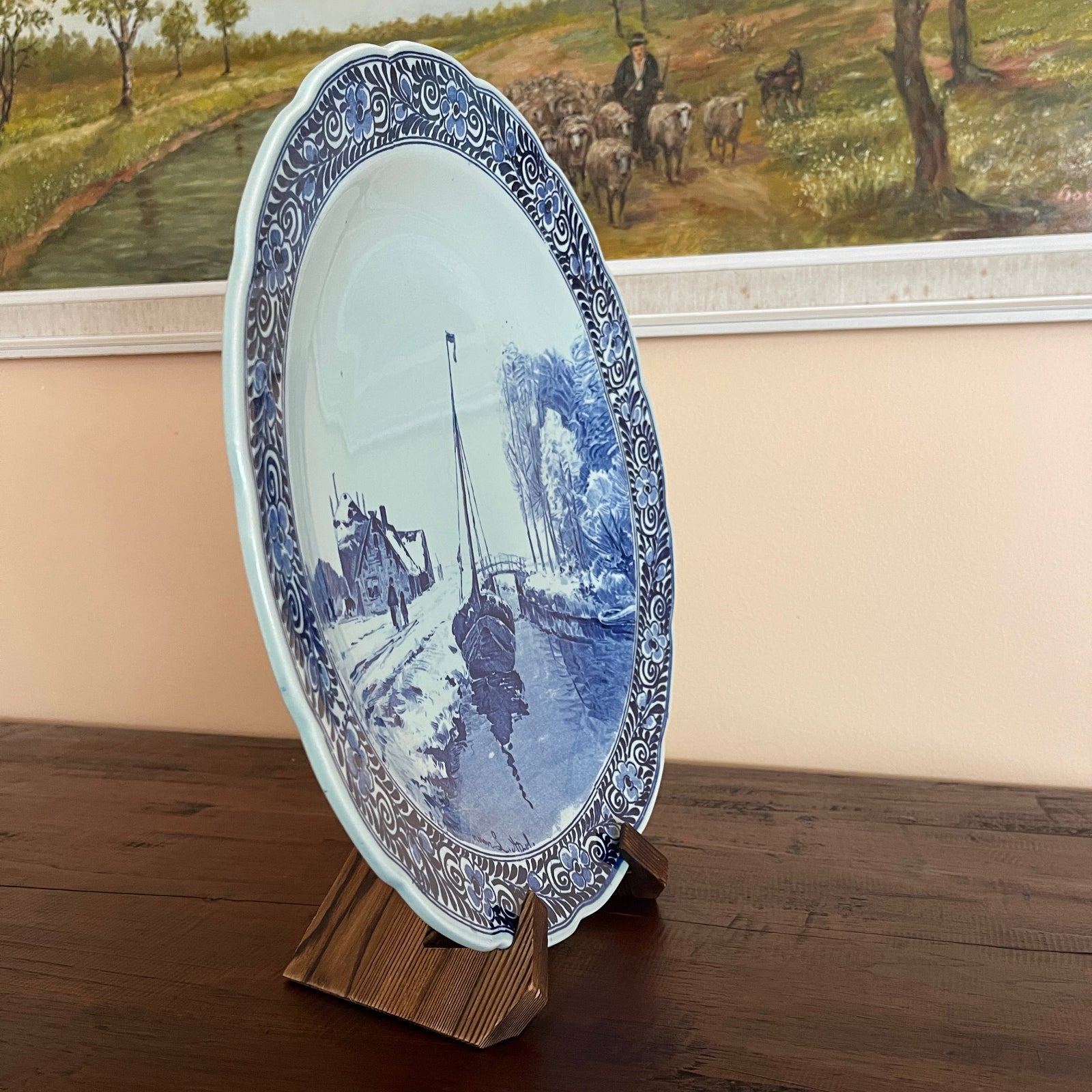 Decorative plate with blue and white design on a wooden stand, with a painting of a pastoral scene in the background.