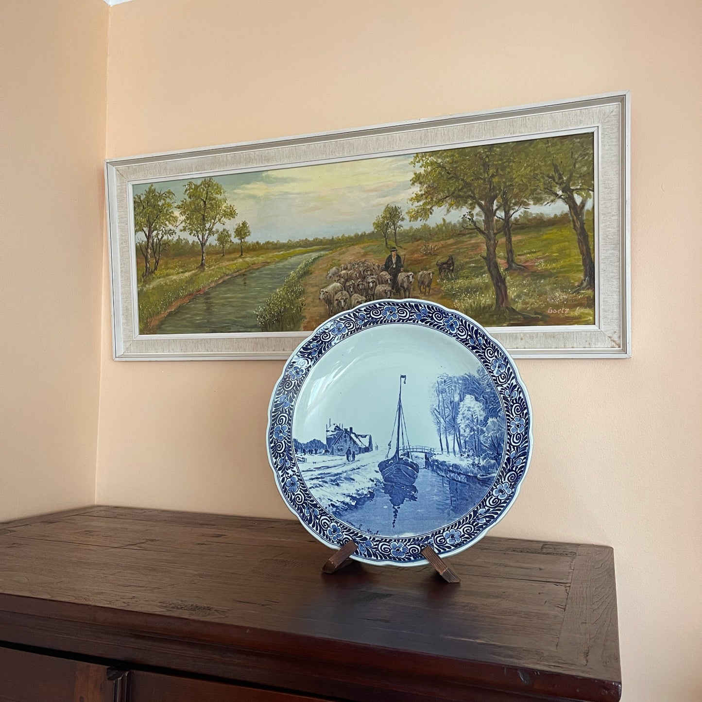 Blue and white decorative plate on a stand with a framed landscape painting in the background.
