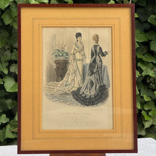 Framed vintage print of two women in elaborate dresses with a decorative border.