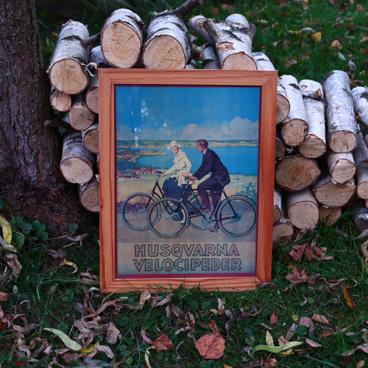 A framed poster depicting two people riding bicycles outdoors, with a wooden frame and the text 'HUSQVARNA VELOCIPEDER' on the lower part.