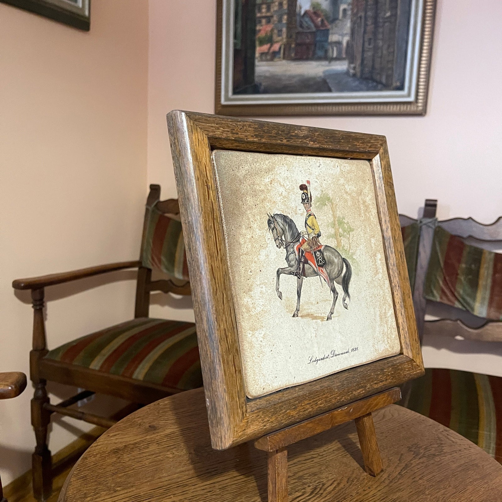 Framed artwork of a person on horseback on a wooden stand with chairs and another framed picture in the background.
