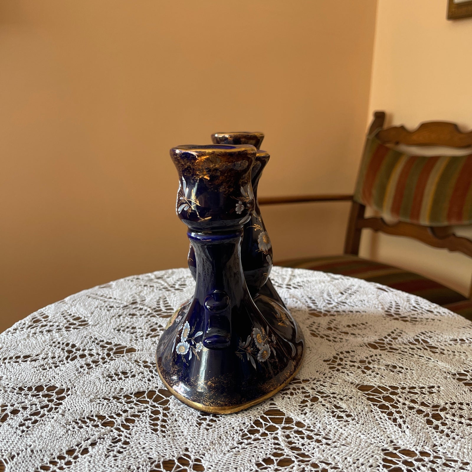 Black ceramic candle holder on a lace tablecloth with a chair and striped cushion in the background.
