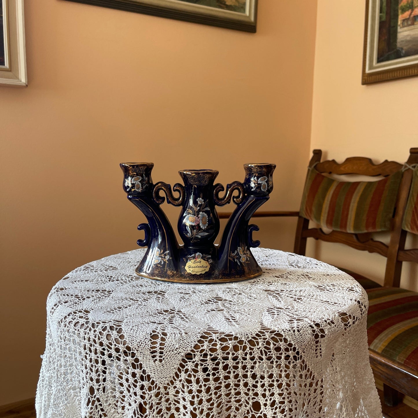 Decorative black metal candle holder on a lace tablecloth with a chair and framed pictures in the background.