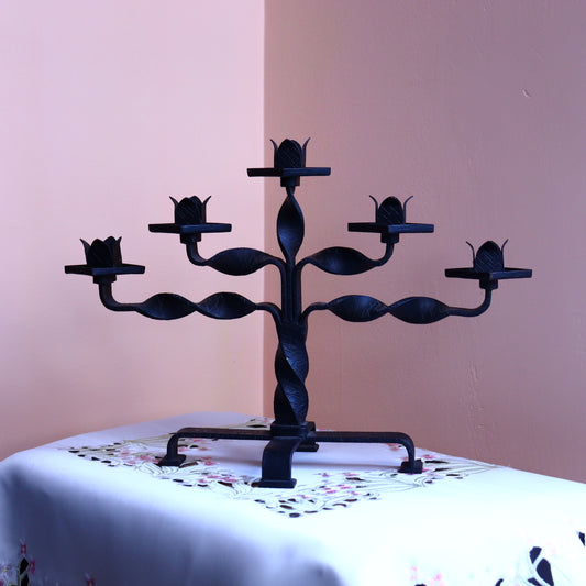 A black five-arm metal candle holder with a decorative floral pattern on the holder and five cups on the arms, placed on a table with a pink background.