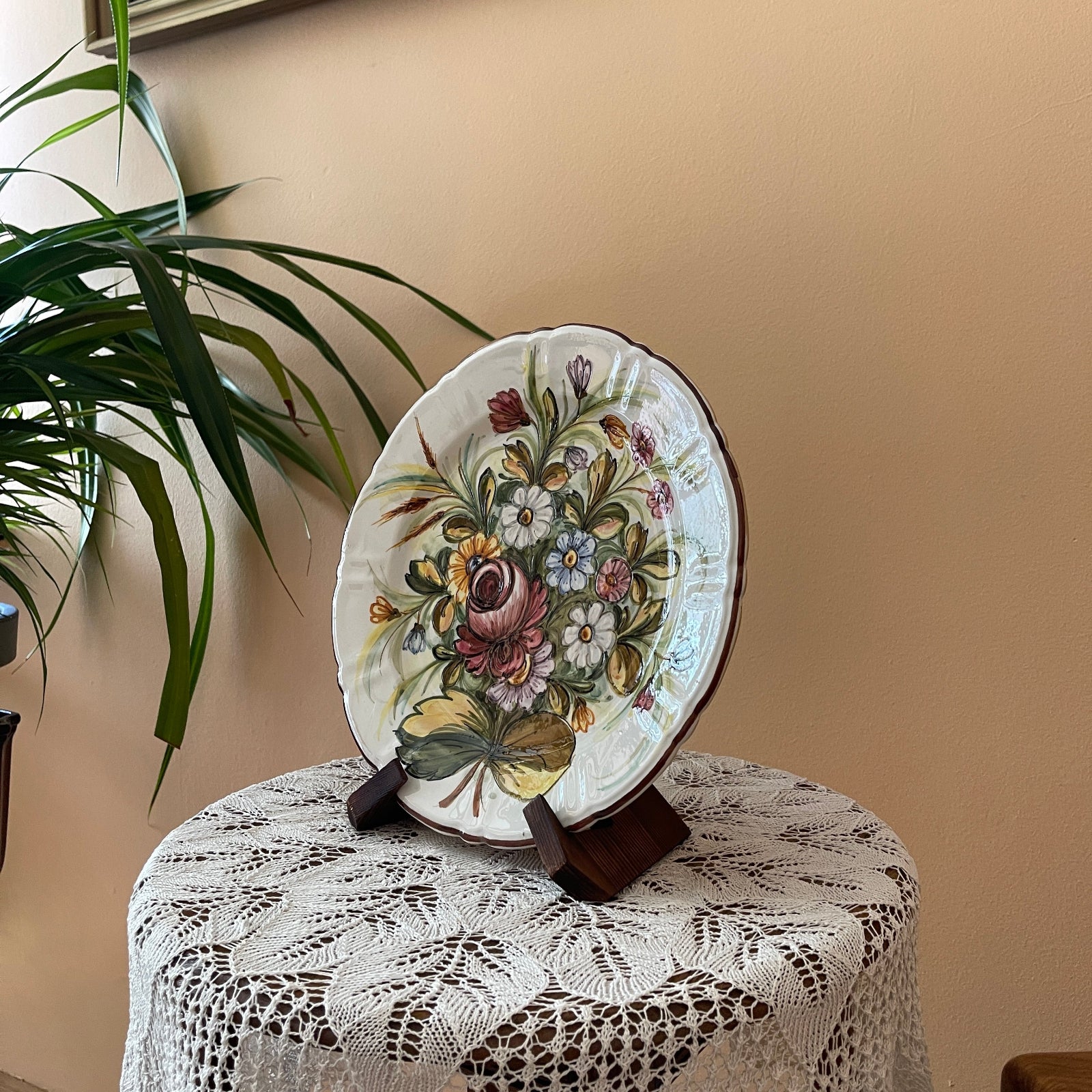 Decorative plate with floral design on a stand against a beige wall with a plant in the corner.