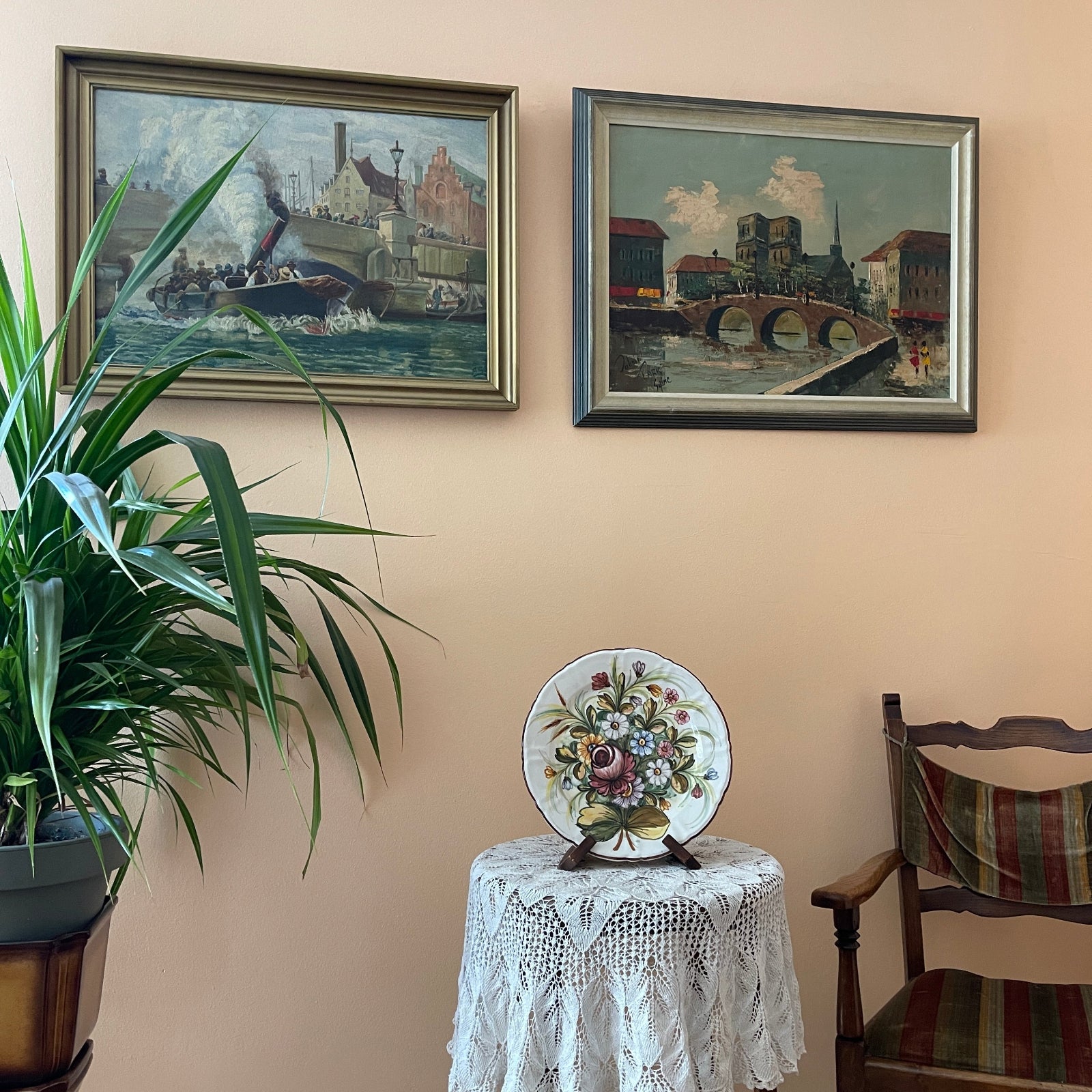 Two framed paintings of a canal scene on a wall above a table with a decorative plate and a plant.