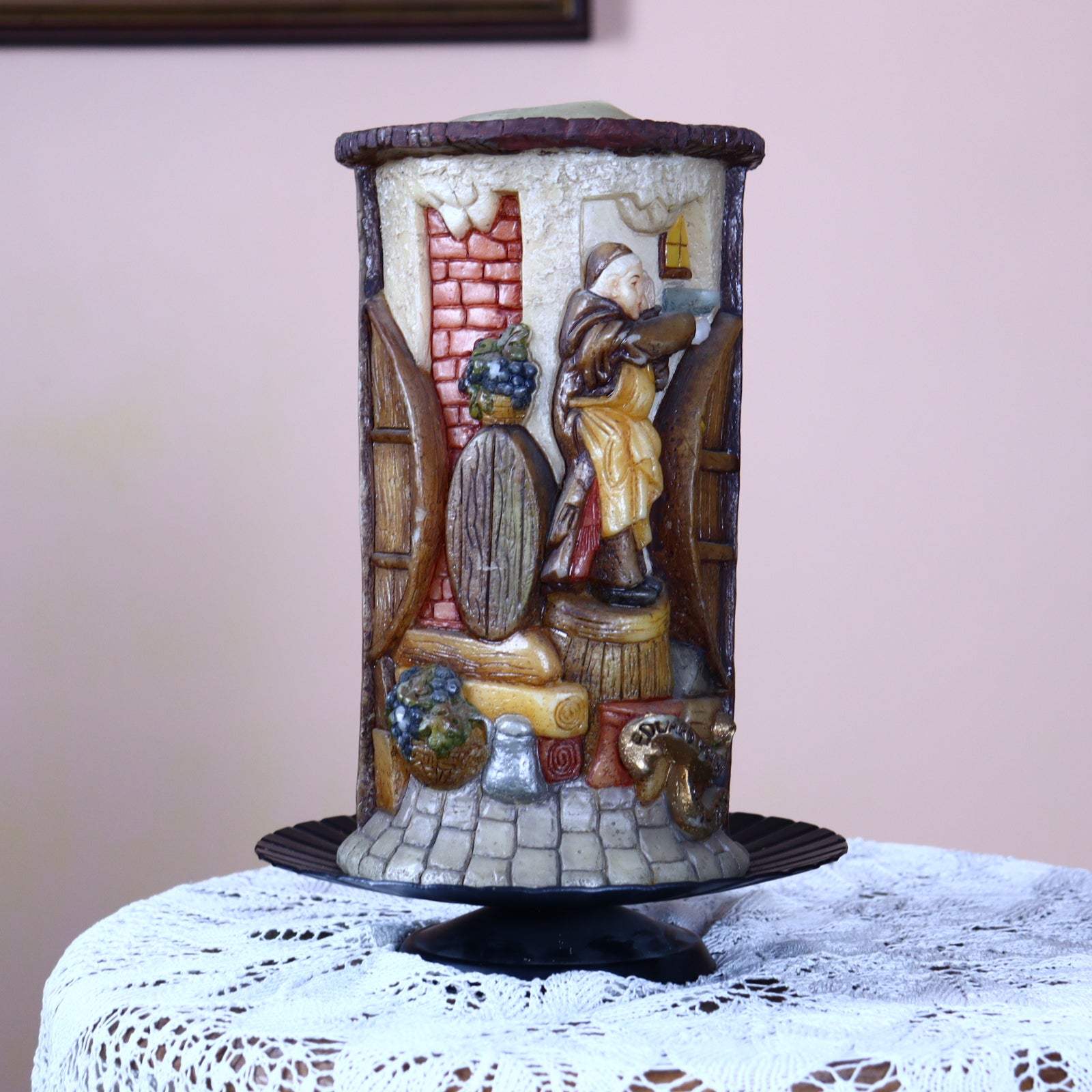 An old hand-made candle decorated with figures of monks, placed on a lace doily, with a painted background mimicking a scene of monks.