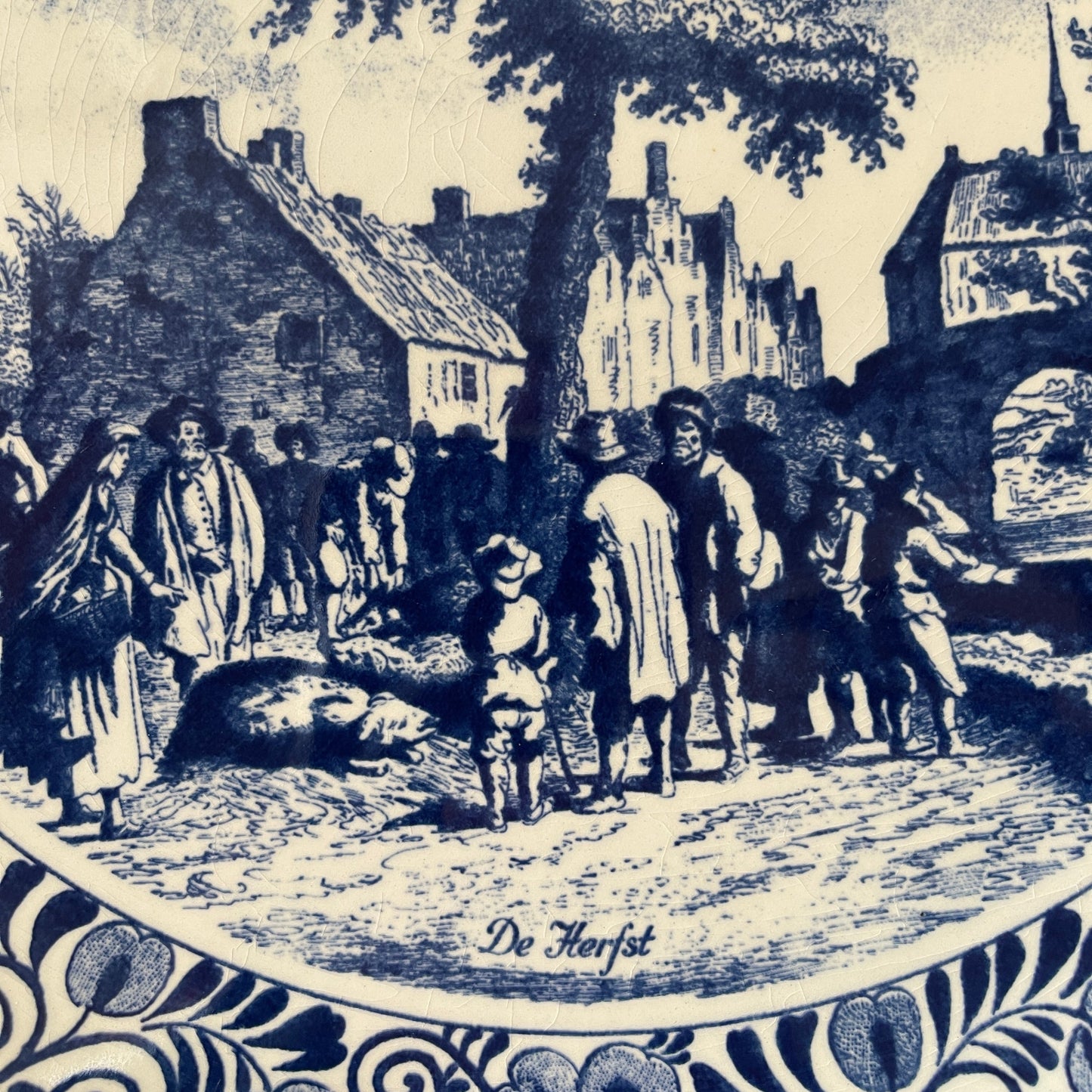 Vintage illustration of a village scene with people and animals, featuring the text 'De Herfst'.