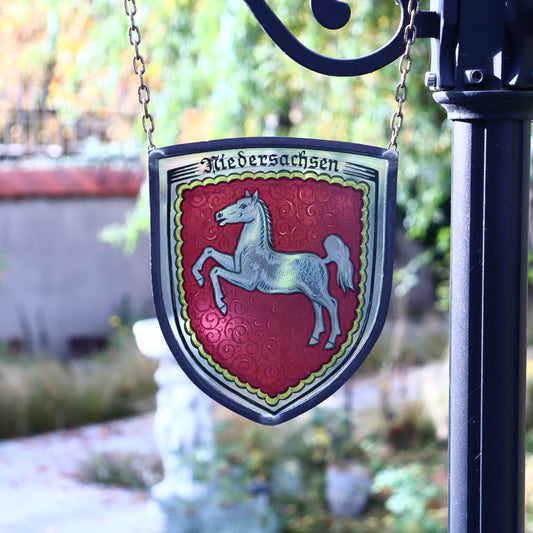 A glass coat of arms of Lower Saxony, Germany, featuring a red and white emblem with a horse on it, framed in lead with a long chain, mounted on a black metal pole.