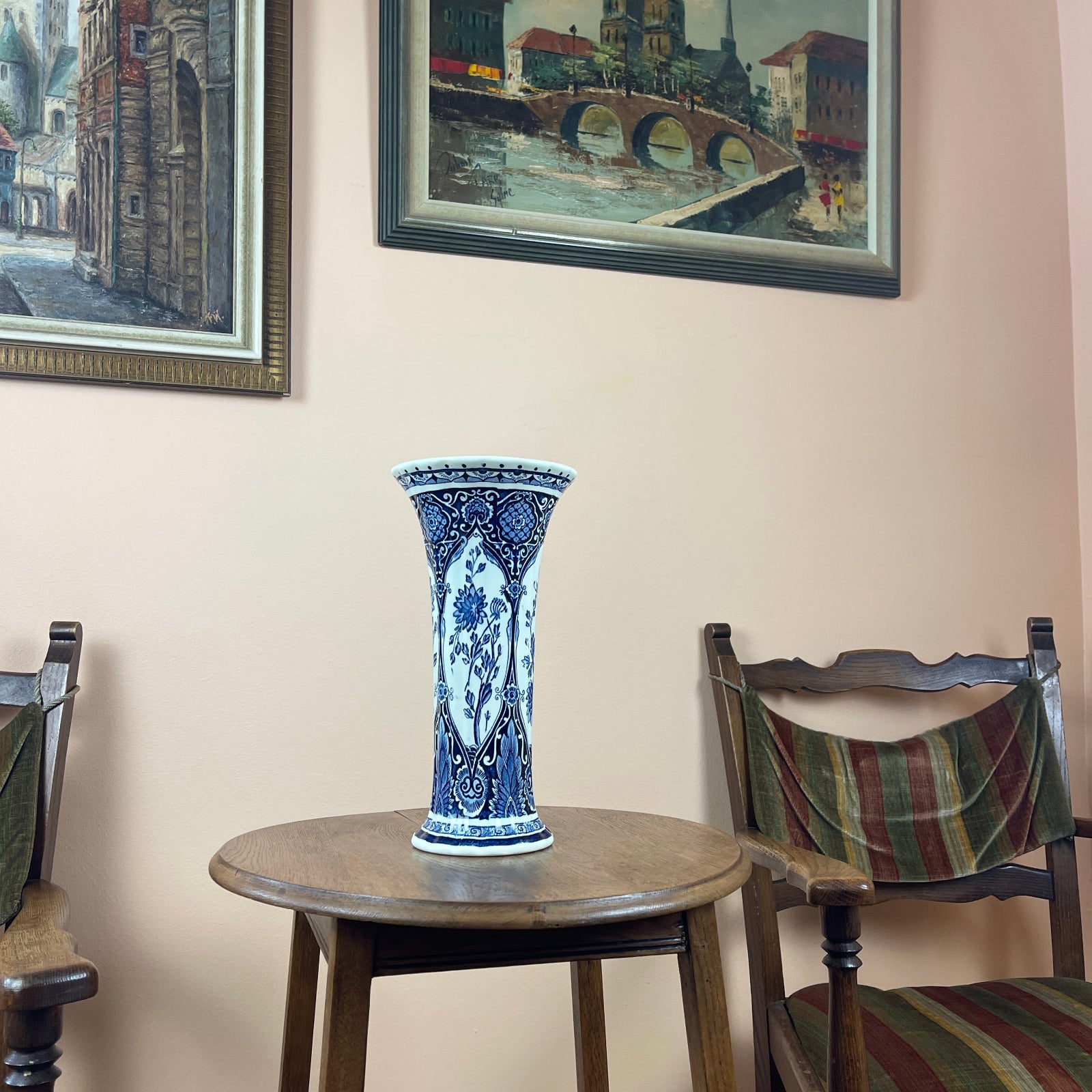 Blue and white vase on a wooden table with two chairs and framed pictures in the background.