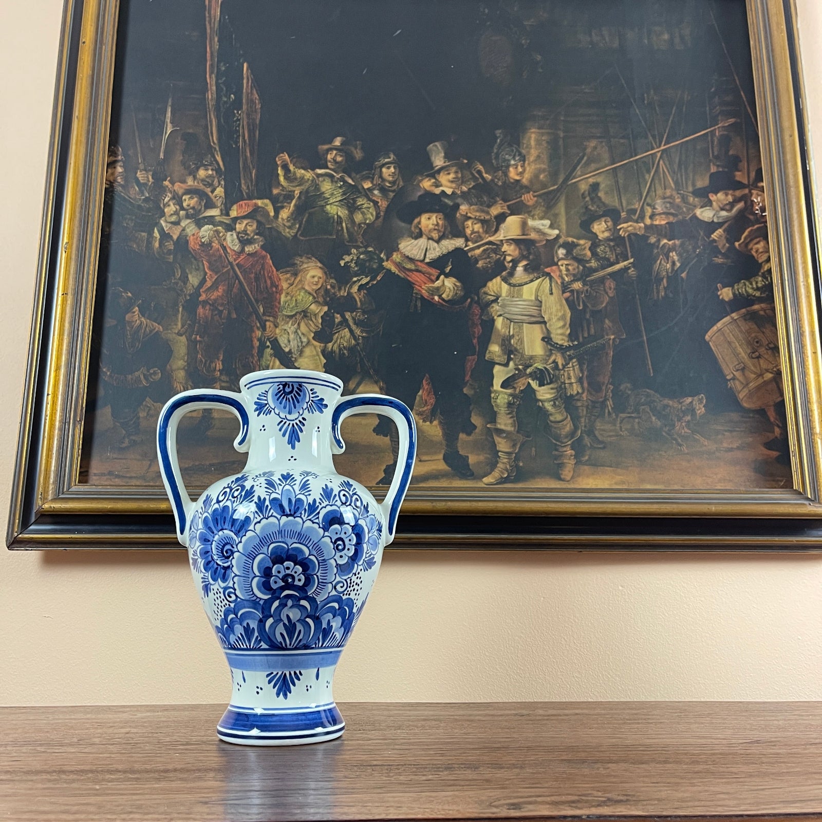 Blue and white ceramic vase on a table with a framed painting of 'The Night Watch' in the background.