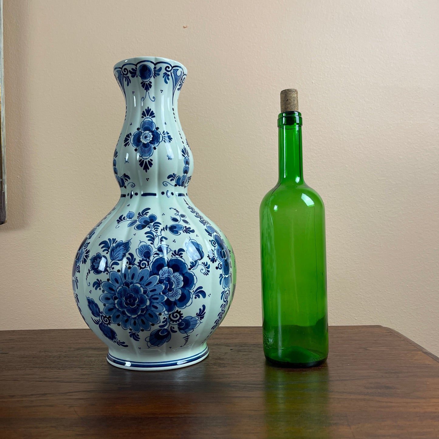 Blue and white floral-patterned vase next to a green bottle on a wooden surface with a beige wall background.