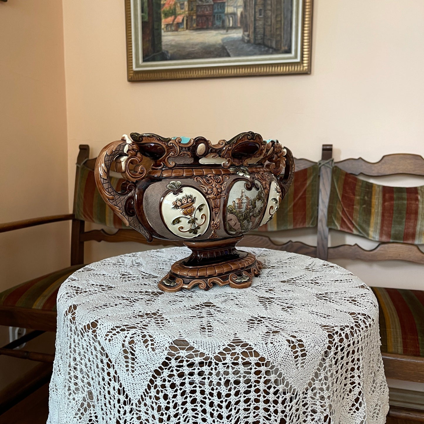 Decorative vase on a table with a lace tablecloth, wooden chairs, and a framed picture in the background.