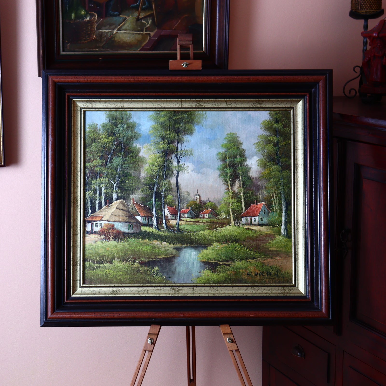 A framed oil painting of cottages in a birch forest, displayed on an easel with a wooden frame, set against a wall with other framed artworks.