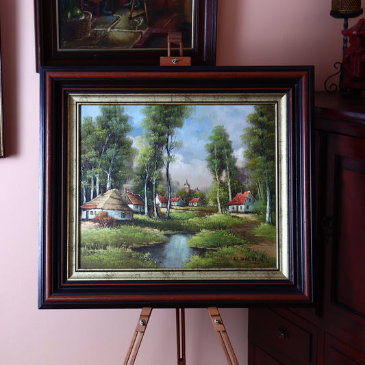 A framed oil painting of cottages in a birch forest, displayed on an easel with a wooden frame, set against a wall with other framed artworks.