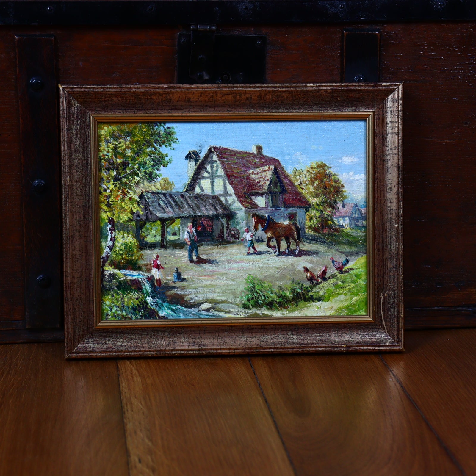 An oil painting of a farm scene with a house, a horse, and people, framed and set against a wooden background.