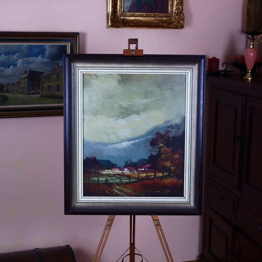 A canvas painting of an autumn meadow with a stormy sky in the background, featuring a village and windmill. The painting is framed and displayed on a easel in a home setting.