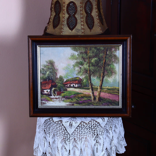 A canvas painting depicting a watermill with a farmhouse in the background, framed in a wooden frame, placed on a lace tablecloth.
