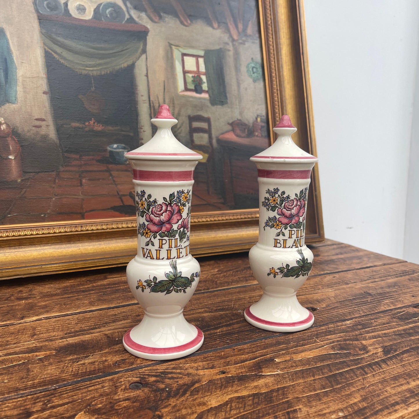 Two decorative urns with floral designs on a wooden surface in front of a framed painting.