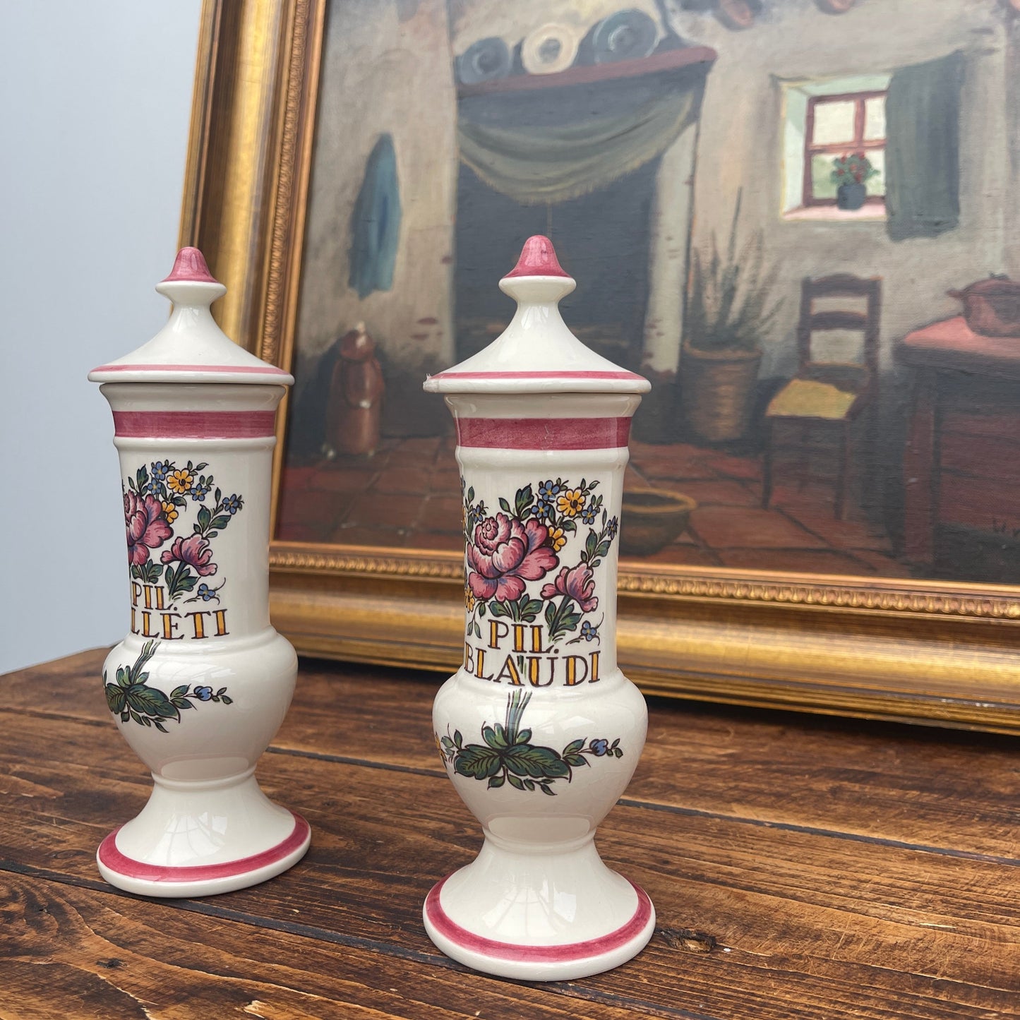 Two decorative vases with floral designs on a wooden surface in front of a framed painting.