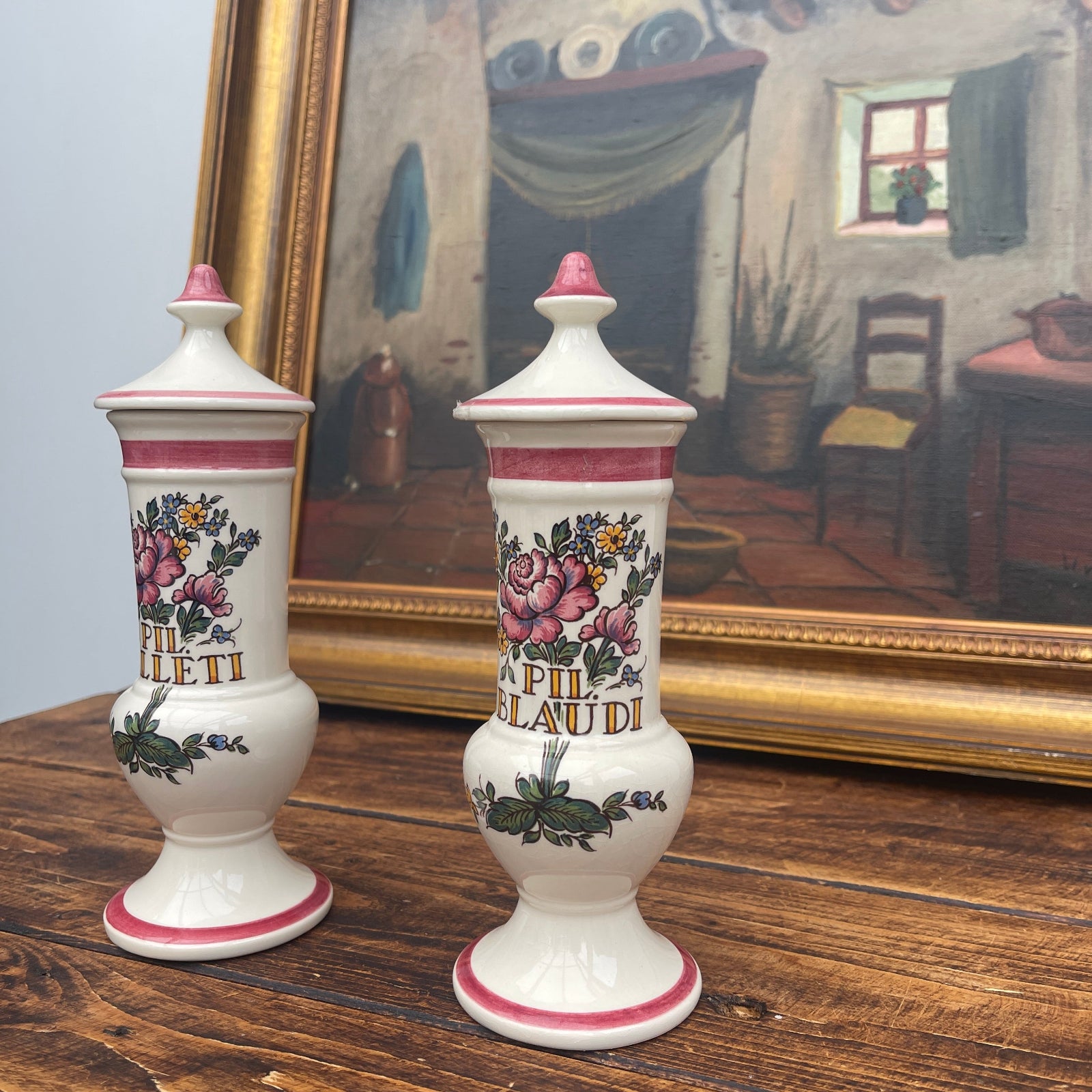 Two decorative vases with floral designs on a wooden surface in front of a framed painting.
