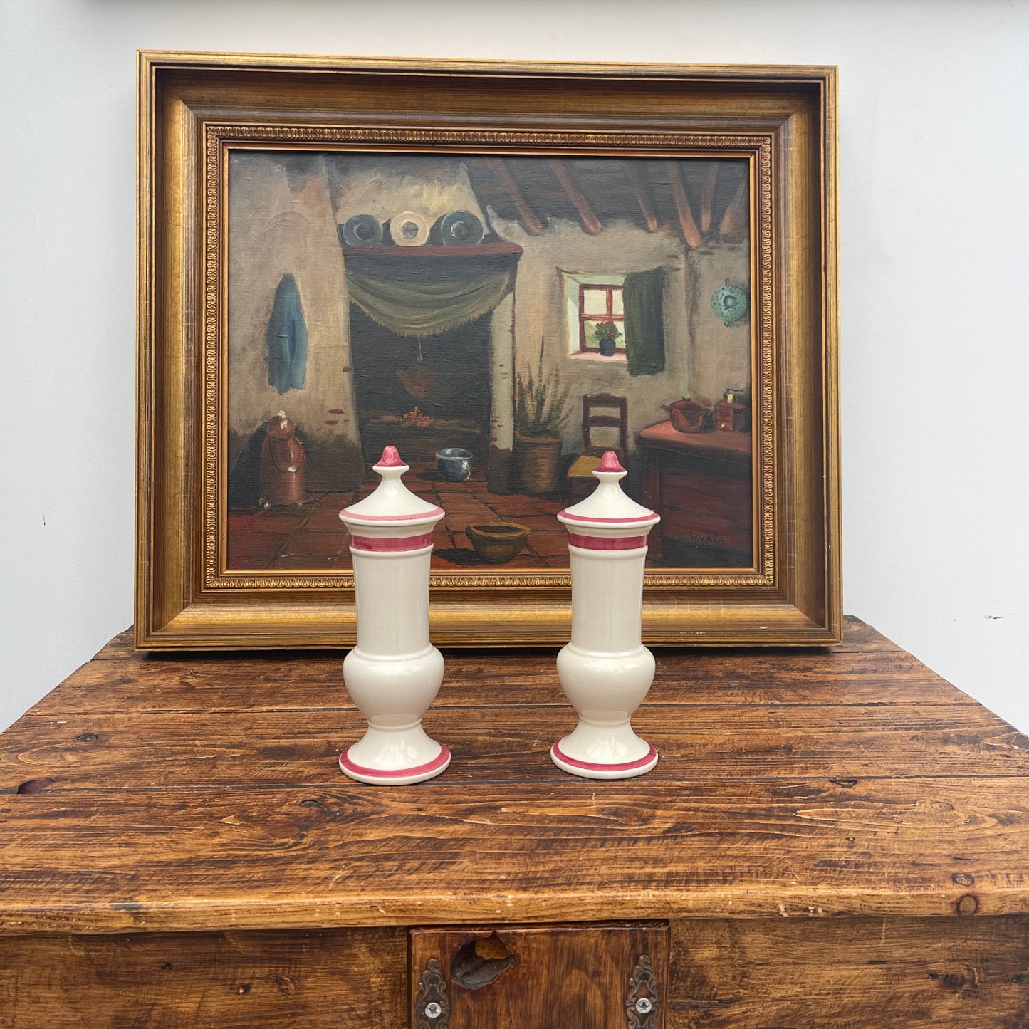 Two white urns with red bands on a wooden surface in front of a framed painting.