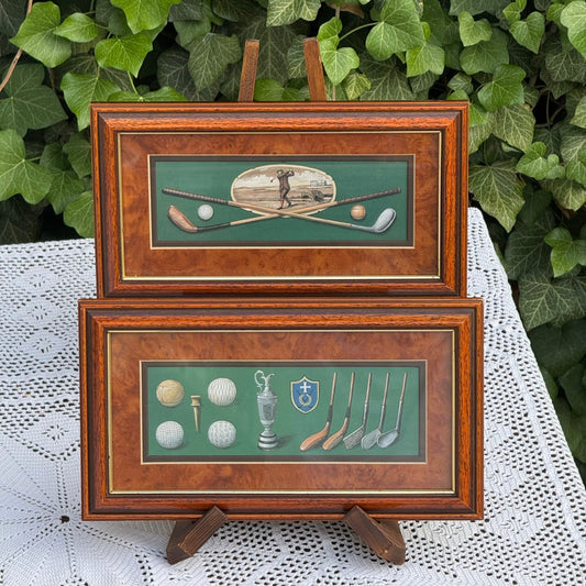Two framed pictures of golf-themed artwork on a white lace tablecloth with green foliage in the background.