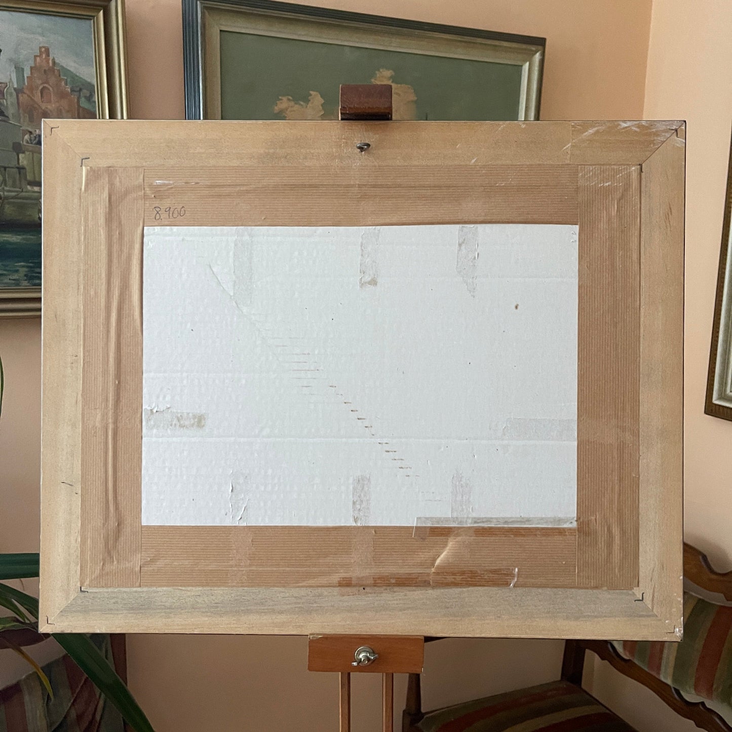 Wooden art easel with a blank white canvas on a stand against a wall with framed pictures.