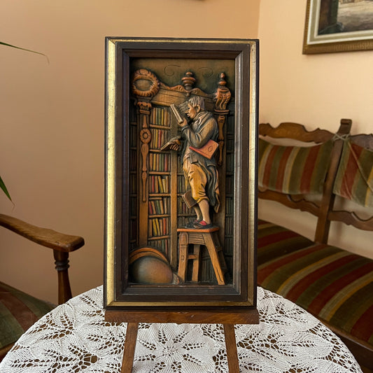 Decorative wall art of a man reading a book in a library setting, framed in a room with a chair and table.