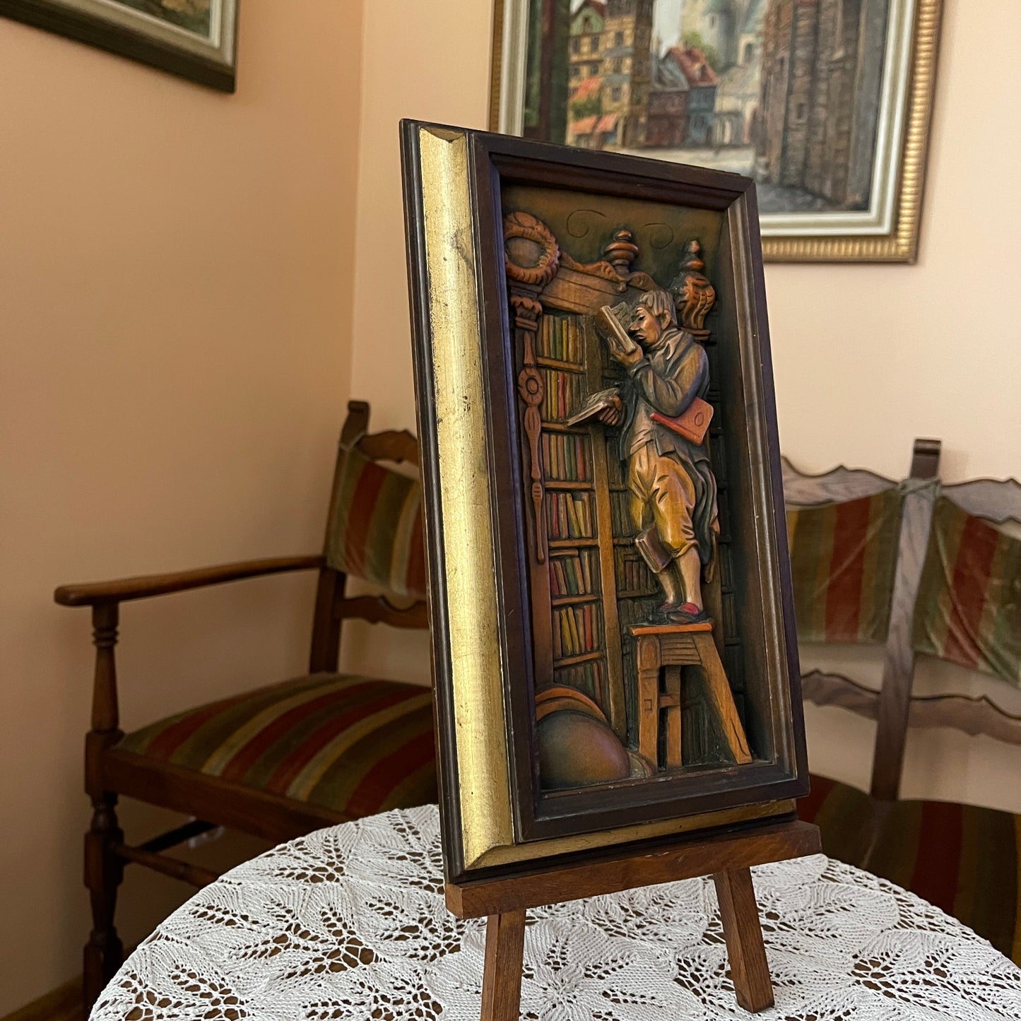 Decorative wall art of a man on a stool with books, framed in a wooden frame, on a table.