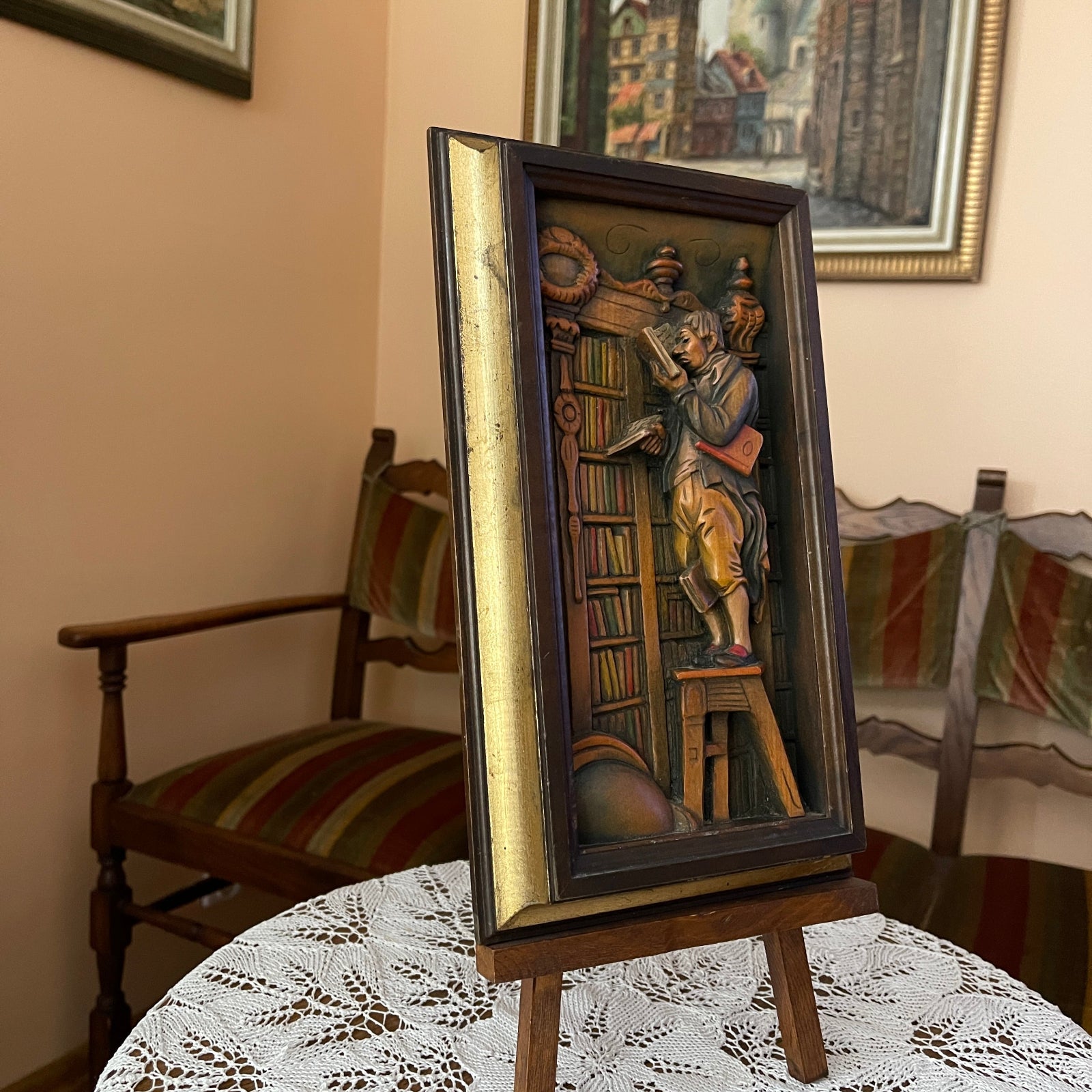 Decorative wall art of a man on a stool with books, framed in a wooden frame, on a table.