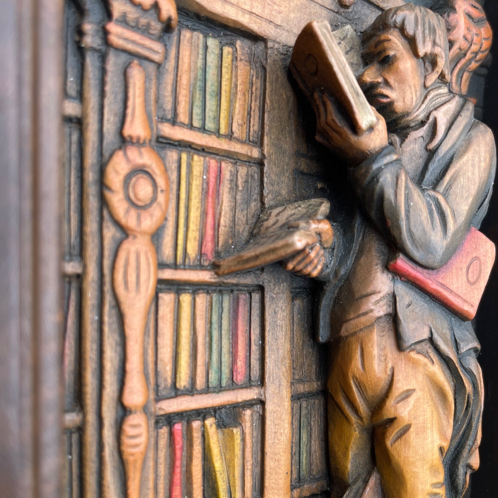 Carved wooden figure of a person reading a book with a bookshelf in the background