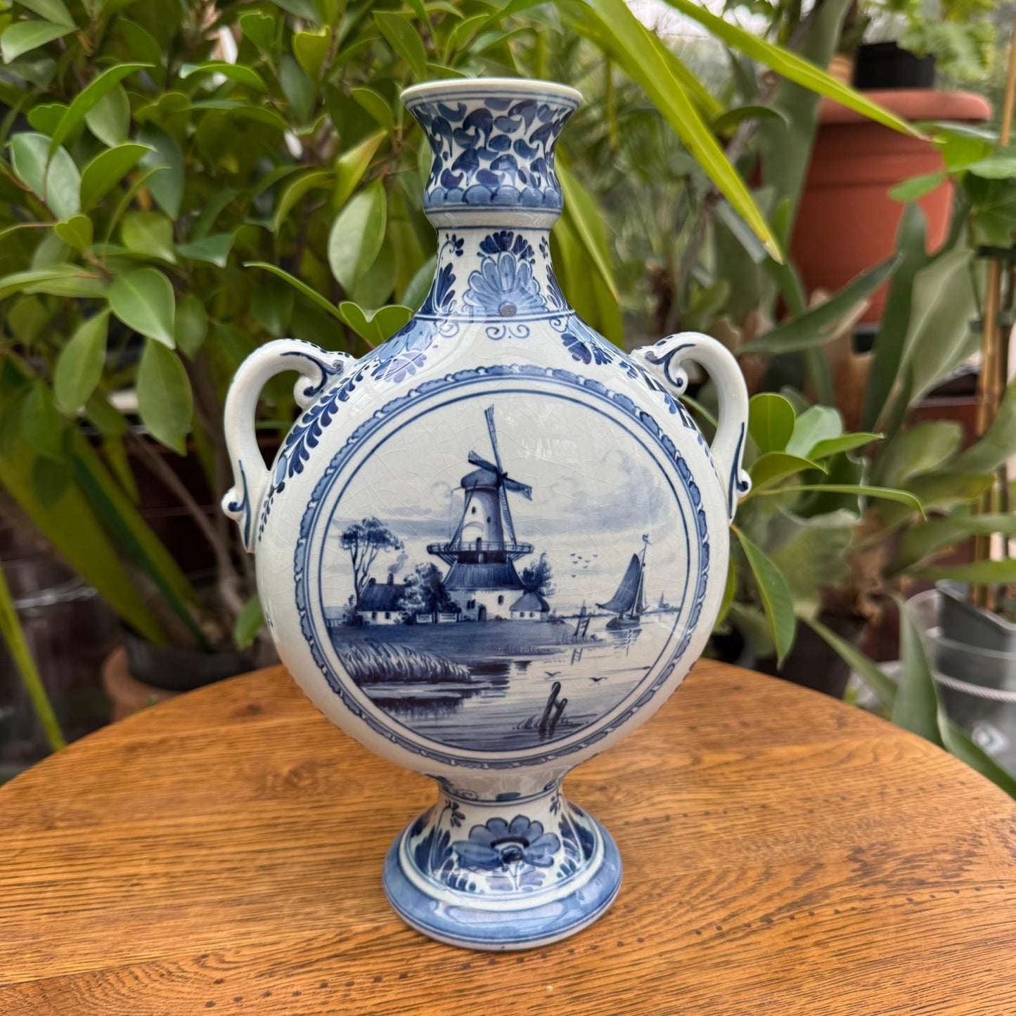 Blue and white ceramic vase with a windmill design on a wooden surface with plants in the background