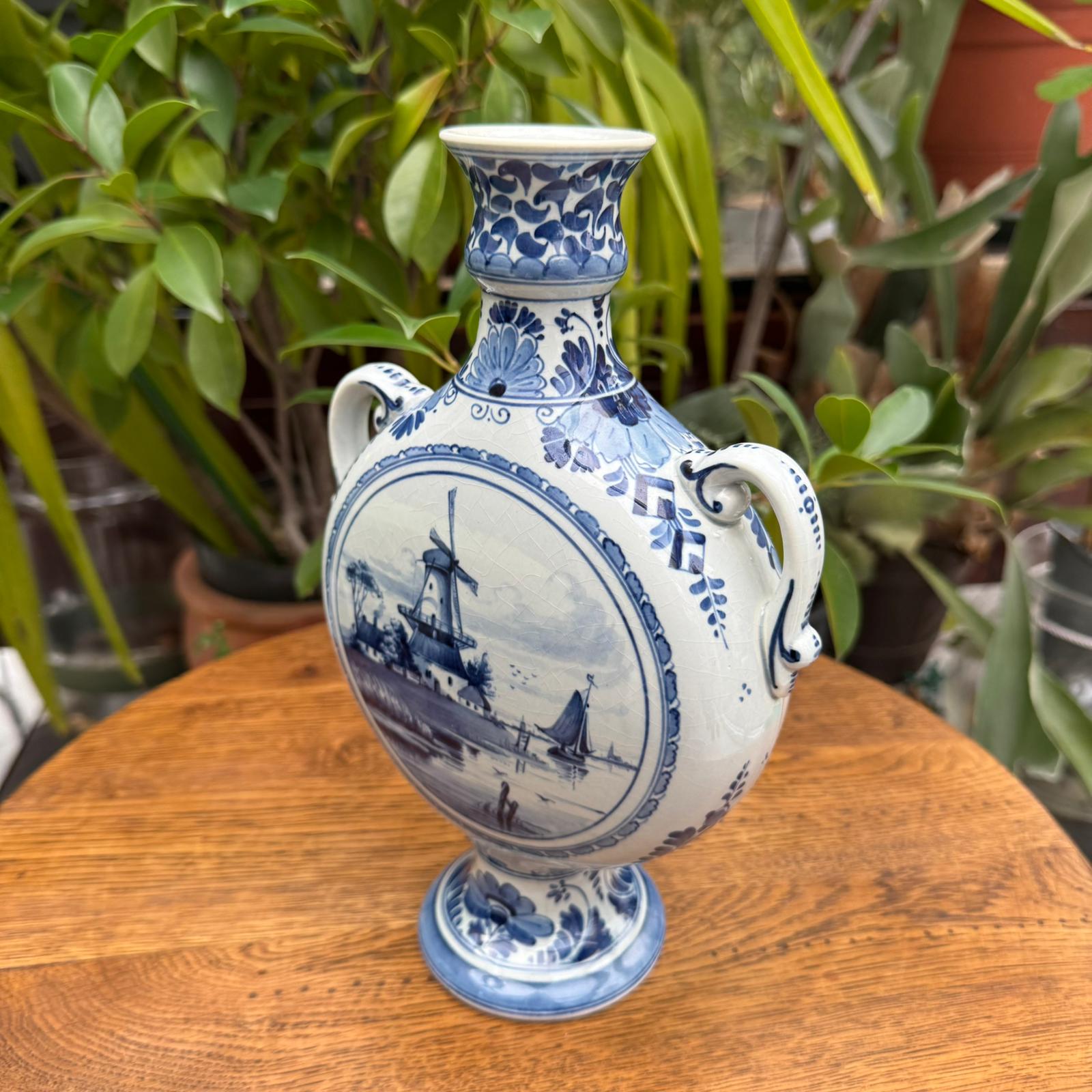 Blue and white decorative vase with a windmill design on a wooden table with plants in the background