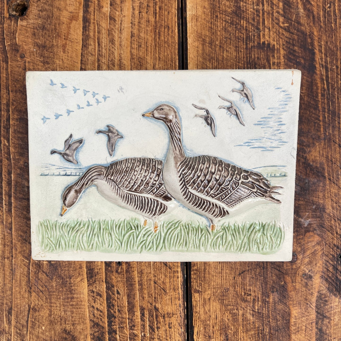 Decorative tile with two geese and flying birds on a wooden surface