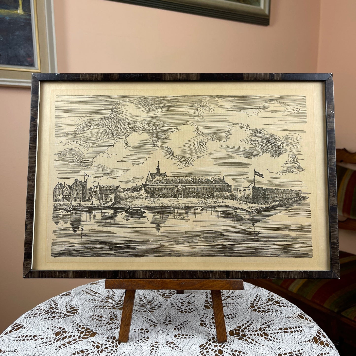 Framed black and white print of a coastal scene on an easel with a lace tablecloth.