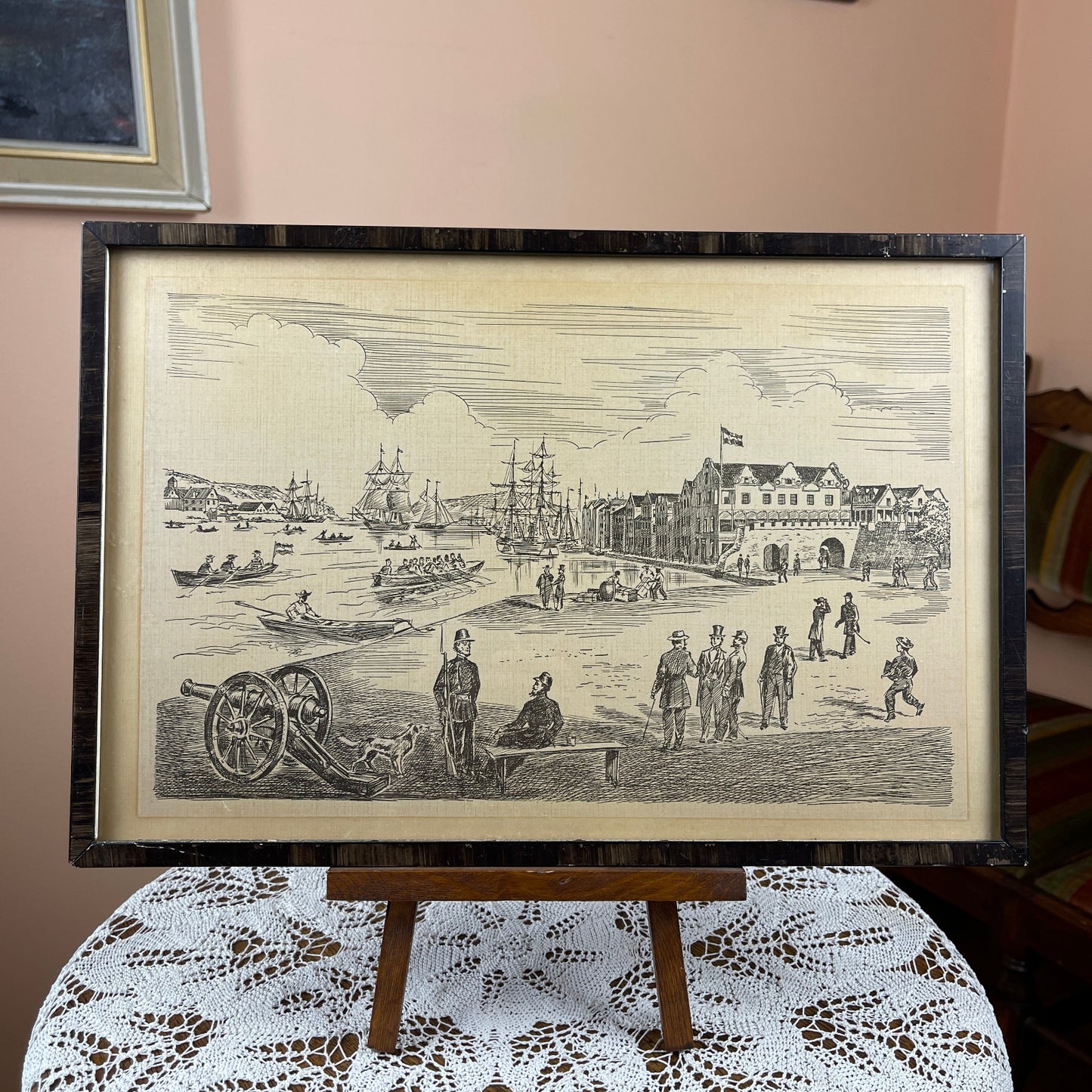 Framed black and white print of a historical scene on an easel with a lace tablecloth.