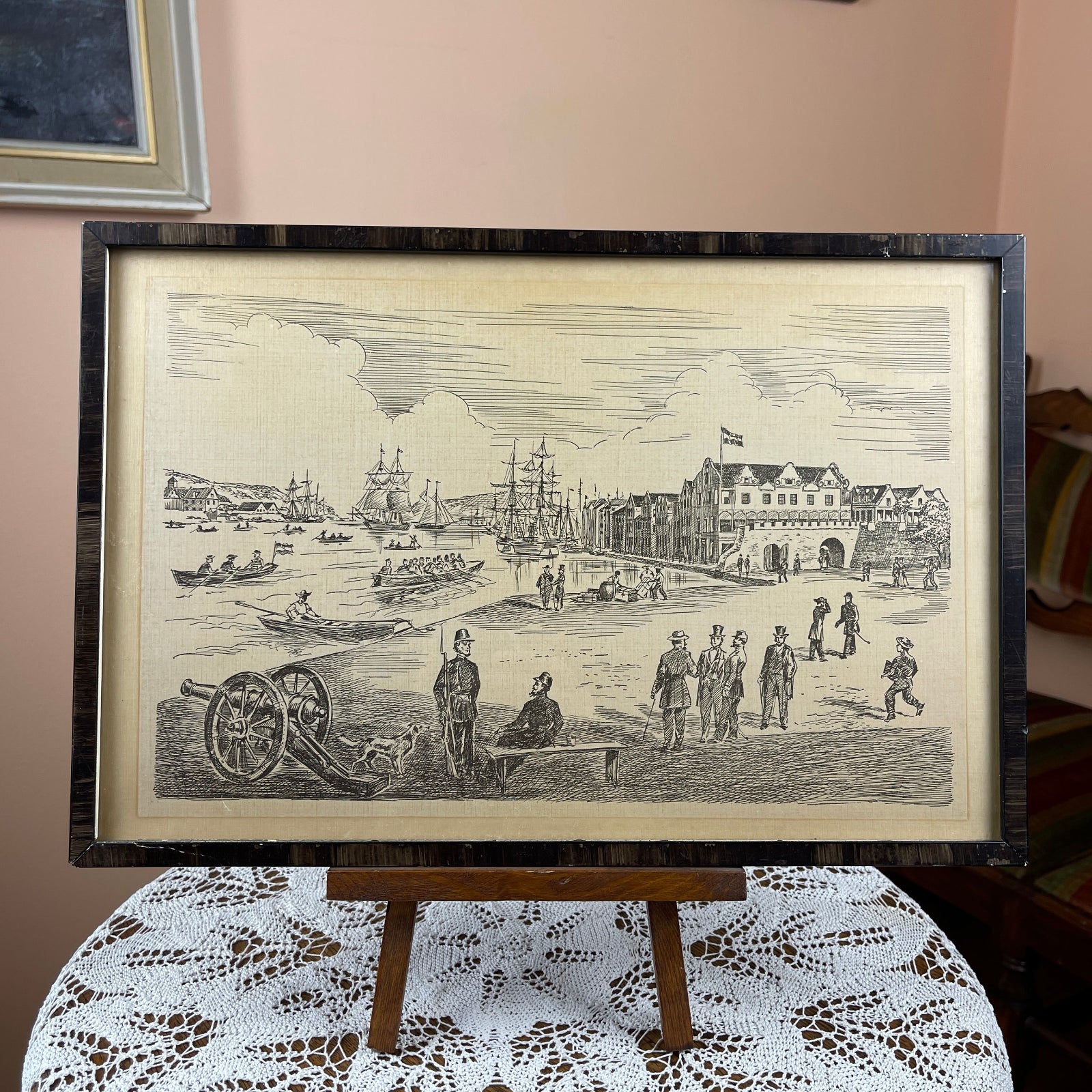 Framed black and white print of a historical scene on an easel with a lace tablecloth.