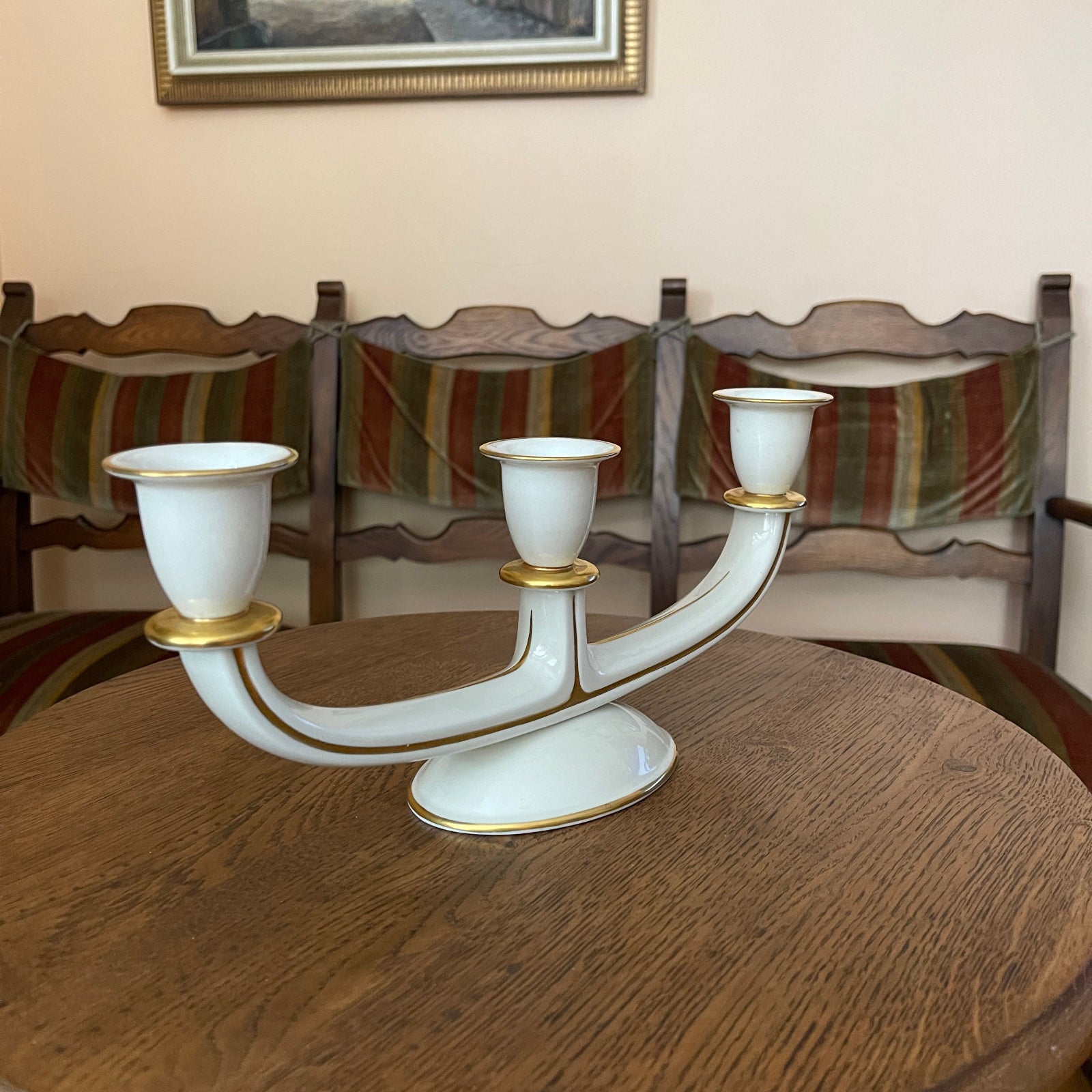 White candelabrum with gold accents on a wooden table, with chairs and a framed picture in the background.
