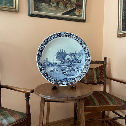 Decorative blue and white plate on a wooden stool with chairs and framed pictures in the background.