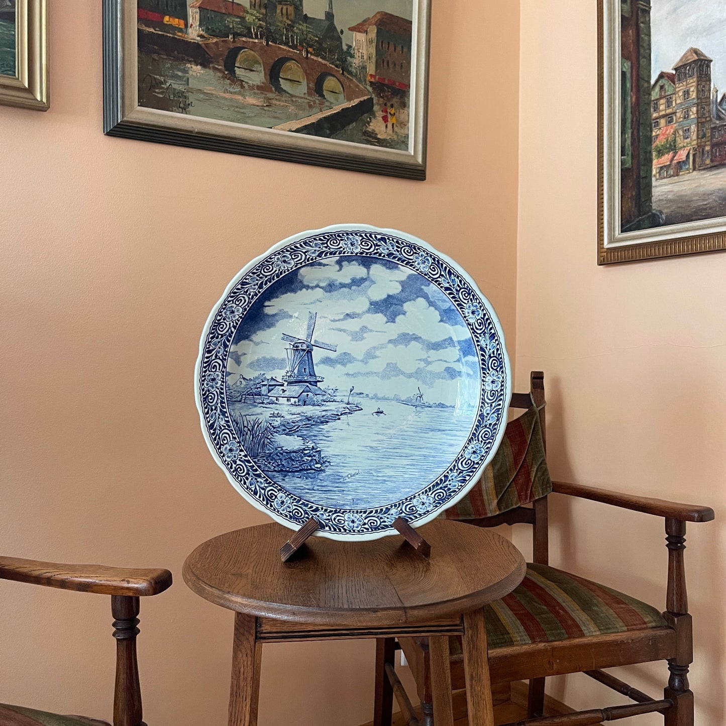 Decorative blue and white plate with a windmill design on a wooden stool against a beige wall with framed pictures.