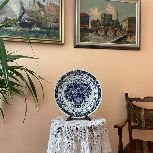 Decorative plate on a stand with two framed paintings on the wall.