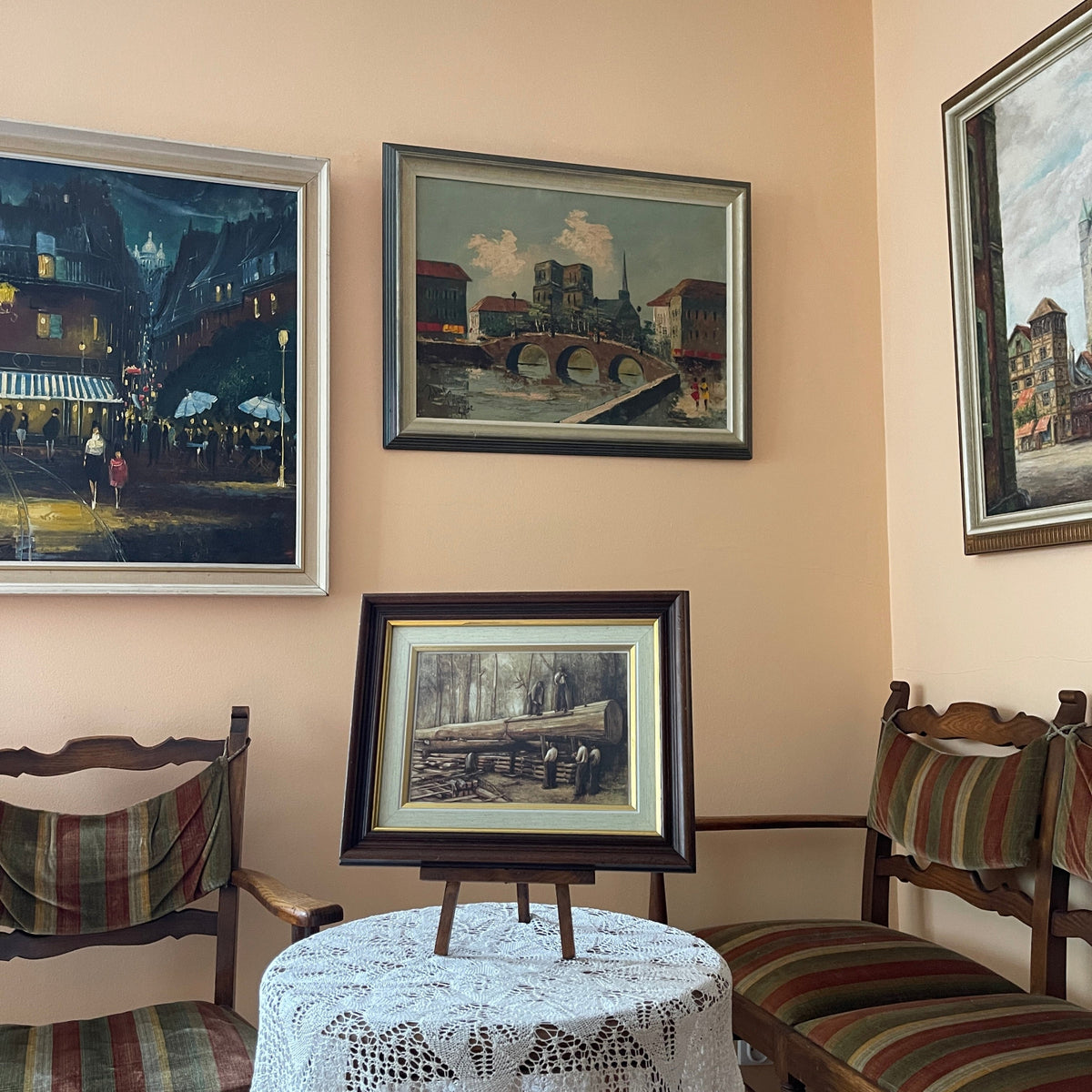 Framed paintings on a wall with chairs and a table in the foreground.