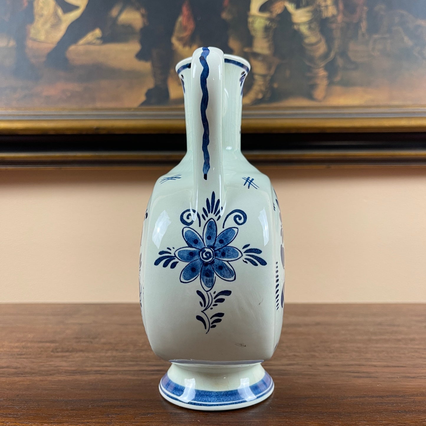 White ceramic vase with blue floral patterns on a wooden surface, with a painting in the background.