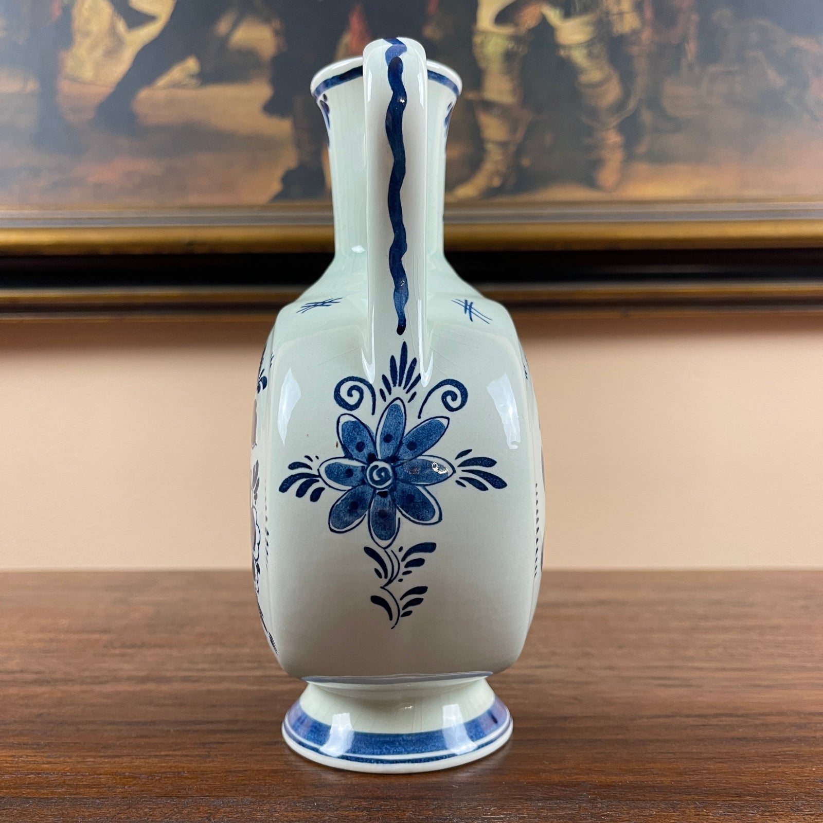 White ceramic vase with blue floral patterns on a wooden surface, with a painting in the background.