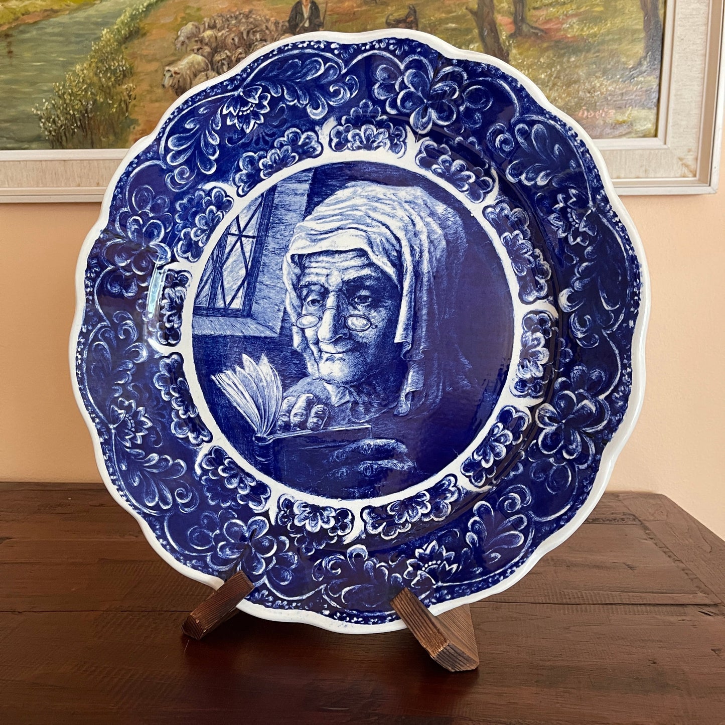 Decorative plate with a blue and white design of an elderly woman reading a book, on a wooden stand.