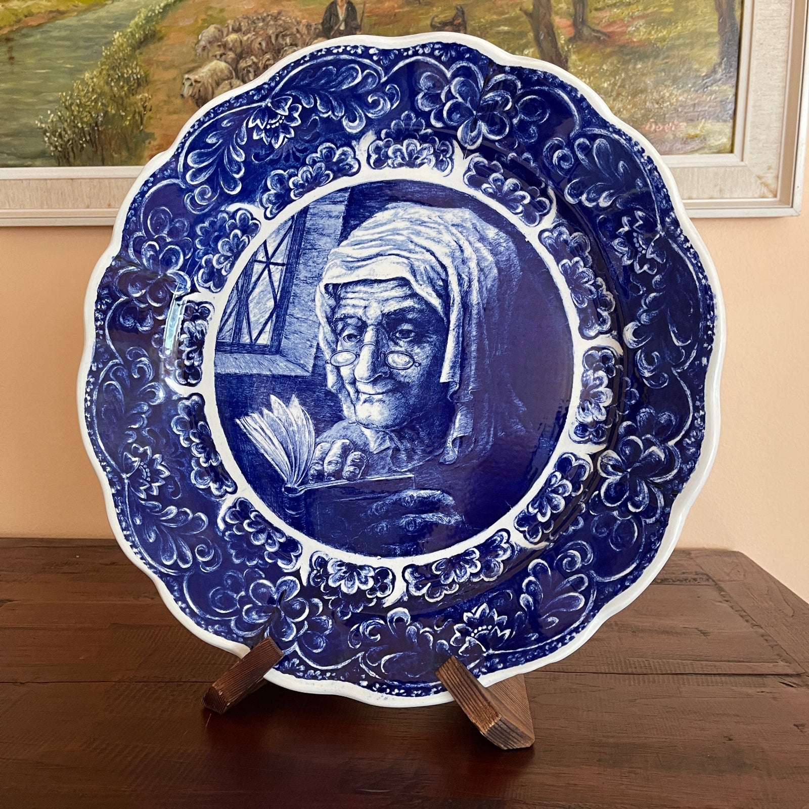Decorative plate with a blue and white design of an elderly woman reading a book, on a wooden stand.